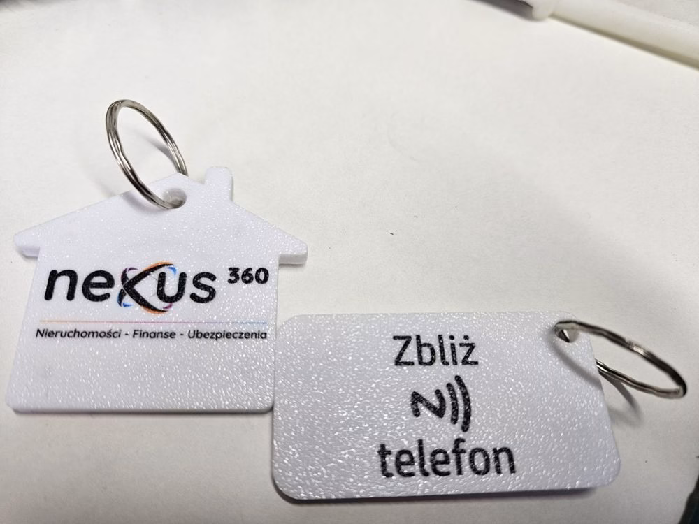 Biały brelok w kształcie domku z logo Nexus360 i drugi z napisem Zbliż telefon