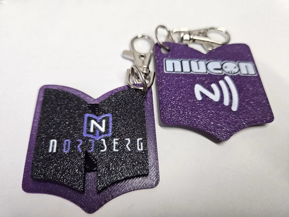 Dwa fioletowe breloki NFC w kształcie otwartej książki z logo Nordberg i Niucon