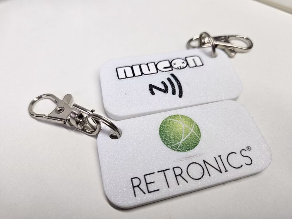 Dwa białe breloki NFC z logo Niucon i Retronics oraz karabińczykami